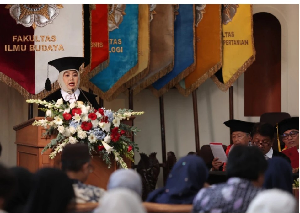 Female graduate in cap and gown speaks at the podium with a large floral arrangement colorful banners behind her